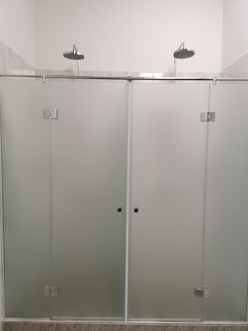 A modern shower setup with frosted glass doors and two overhead showerheads. The setting is minimalistic with clean, straight lines and a focus on functionality.