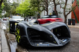 A sleek, futuristic autonomous race car with a matte black finish and neon yellow accents is parked on a street lined with trees. The road appears wet, suggesting recent rain. In the background, several other vehicles, including a blurred red car in motion, are visible, along with street signs and banners on nearby buildings.