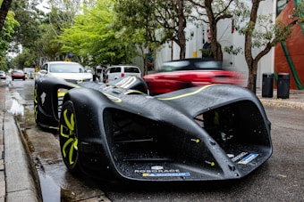 A sleek, futuristic autonomous race car with a matte black finish and neon yellow accents is parked on a street lined with trees. The road appears wet, suggesting recent rain. In the background, several other vehicles, including a blurred red car in motion, are visible, along with street signs and banners on nearby buildings.