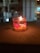 A small lit candle in a transparent glass container, placed on a flat surface. The candle is surrounded by a translucent gel with embedded decorative objects, including pink and yellow elements. The background is blurred, featuring some indistinct lights and architectural structures.
