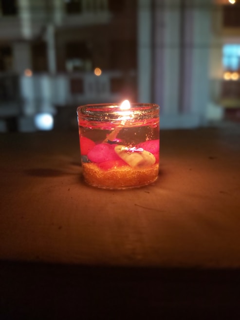 A small lit candle in a transparent glass container, placed on a flat surface. The candle is surrounded by a translucent gel with embedded decorative objects, including pink and yellow elements. The background is blurred, featuring some indistinct lights and architectural structures.