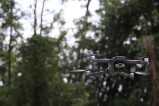 A drone is captured in mid-flight, surrounded by a backdrop of lush green foliage. The drone is dark gray with rotors spinning and a camera attached to its front.