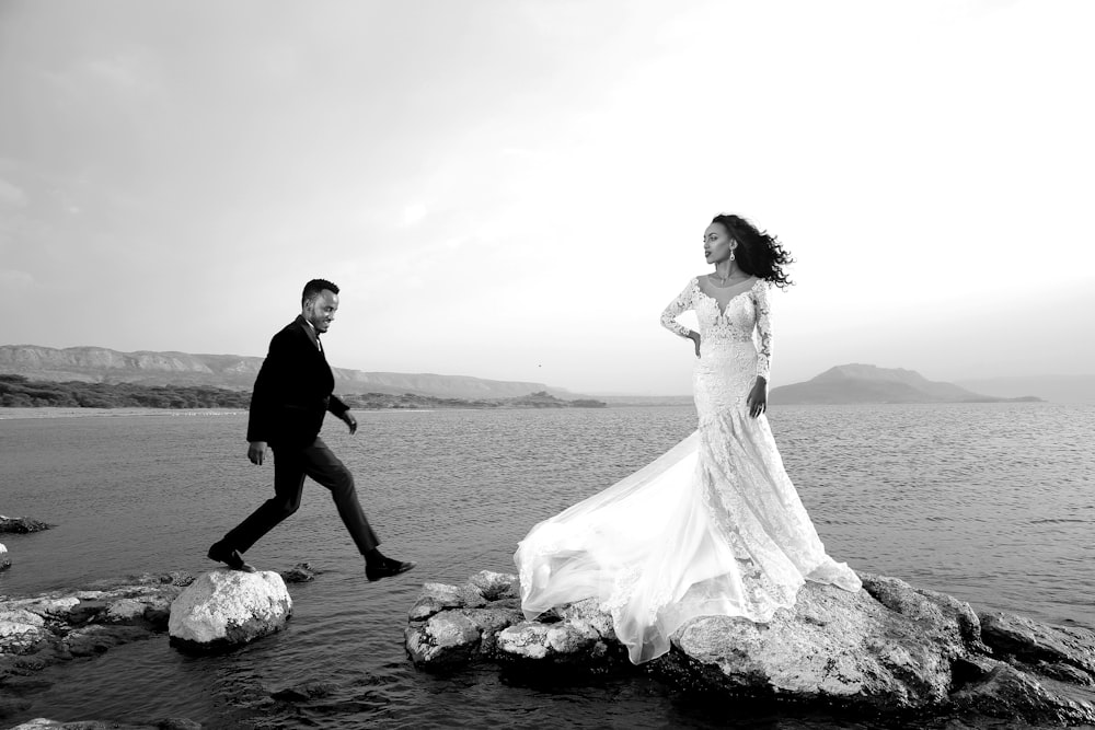 Foto en escala de grises de la novia y el novio de pie en una roca cerca del cuerpo de agua