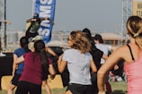 A group of people appears to be participating in an outdoor fitness class or dance session. They are dressed in athletic clothing, and a blue banner can be seen in the background, along with a stage. The setting suggests a lively and energetic atmosphere.