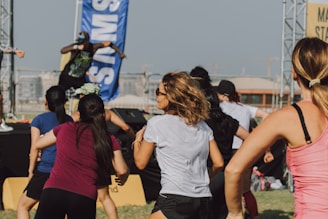 A group of people appears to be participating in an outdoor fitness class or dance session. They are dressed in athletic clothing, and a blue banner can be seen in the background, along with a stage. The setting suggests a lively and energetic atmosphere.