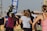 A group of people appears to be participating in an outdoor fitness class or dance session. They are dressed in athletic clothing, and a blue banner can be seen in the background, along with a stage. The setting suggests a lively and energetic atmosphere.