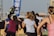 A group of people appears to be participating in an outdoor fitness class or dance session. They are dressed in athletic clothing, and a blue banner can be seen in the background, along with a stage. The setting suggests a lively and energetic atmosphere.