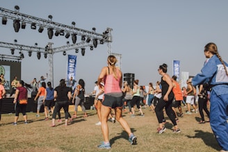 A group of people is actively participating in an outdoor exercise class. They are standing on a grassy field in front of a stage equipped with lighting and speakers. Most participants are wearing athletic clothing and appear to be following the instructors on stage who are guiding the workout.