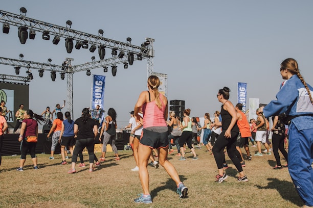 A group of people is actively participating in an outdoor exercise class. They are standing on a grassy field in front of a stage equipped with lighting and speakers. Most participants are wearing athletic clothing and appear to be following the instructors on stage who are guiding the workout.