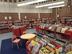 A well-organized children's section of a library features red shelving units filled with colorful children's books. The room is spacious with a high ceiling and large windows. There are small tables and chairs placed on a blue carpet, creating a cozy reading area. The walls are lined with brick and the lighting is bright, giving the space a welcoming atmosphere.