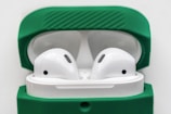 A pair of wireless earbuds resting inside an open charging case with a green cover. The earbuds are white, featuring a sleek and modern design, and the green cover adds a vibrant contrast.