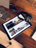 A wooden table holds a tablet displaying a streaming service interface with various movie and TV show thumbnails. Nearby, a pair of Bose headphones rests on the table along with a keyboard and a mouse.