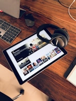A wooden table holds a tablet displaying a streaming service interface with various movie and TV show thumbnails. Nearby, a pair of Bose headphones rests on the table along with a keyboard and a mouse.