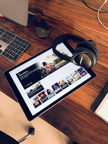 A wooden table holds a tablet displaying a streaming service interface with various movie and TV show thumbnails. Nearby, a pair of Bose headphones rests on the table along with a keyboard and a mouse.