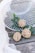Three homemade cookies with chocolate chips rest on top of green leaves, placed on a wooden surface. A white mesh basket is tipped over, adding a rustic touch to the display. A gauzy white fabric drapes softly in the corner.