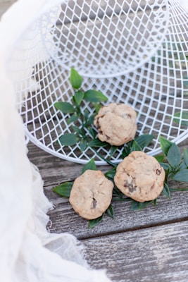 Three homemade cookies with chocolate chips rest on top of green leaves, placed on a wooden surface. A white mesh basket is tipped over, adding a rustic touch to the display. A gauzy white fabric drapes softly in the corner.