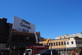 A large billboard with the phrase 'Make Google do it' is prominently displayed above urban buildings. The sky is clear and blue, providing a bright backdrop. Below, various city buildings with businesses and a traffic light are visible, along with a few vehicles and people on the street.