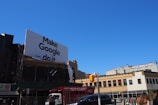 A large billboard with the phrase 'Make Google do it' is prominently displayed above urban buildings. The sky is clear and blue, providing a bright backdrop. Below, various city buildings with businesses and a traffic light are visible, along with a few vehicles and people on the street.