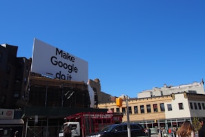 A large billboard with the phrase 'Make Google do it' is prominently displayed above urban buildings. The sky is clear and blue, providing a bright backdrop. Below, various city buildings with businesses and a traffic light are visible, along with a few vehicles and people on the street.