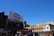 A large billboard with the phrase 'Make Google do it' is prominently displayed above urban buildings. The sky is clear and blue, providing a bright backdrop. Below, various city buildings with businesses and a traffic light are visible, along with a few vehicles and people on the street.