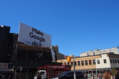 A large billboard with the phrase 'Make Google do it' is prominently displayed above urban buildings. The sky is clear and blue, providing a bright backdrop. Below, various city buildings with businesses and a traffic light are visible, along with a few vehicles and people on the street.