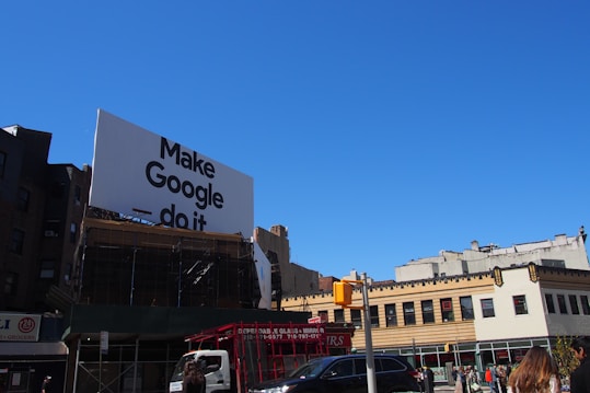 A large billboard with the phrase 'Make Google do it' is prominently displayed above urban buildings. The sky is clear and blue, providing a bright backdrop. Below, various city buildings with businesses and a traffic light are visible, along with a few vehicles and people on the street.