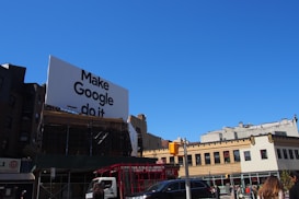 A large billboard with the phrase 'Make Google do it' is prominently displayed above urban buildings. The sky is clear and blue, providing a bright backdrop. Below, various city buildings with businesses and a traffic light are visible, along with a few vehicles and people on the street.