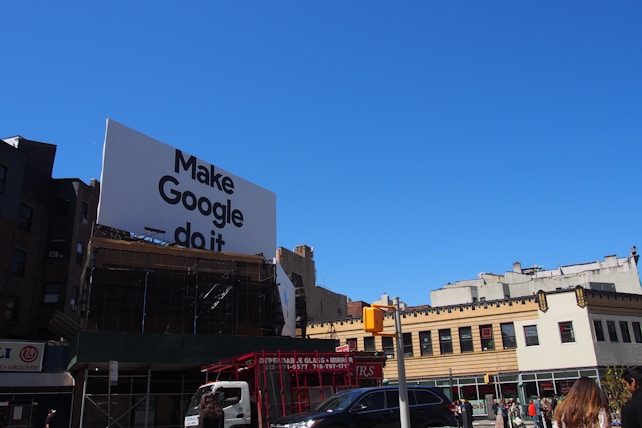 A large billboard with the phrase 'Make Google do it' is prominently displayed above urban buildings. The sky is clear and blue, providing a bright backdrop. Below, various city buildings with businesses and a traffic light are visible, along with a few vehicles and people on the street.