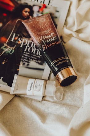A magazine lies on a textured fabric surface, featuring a woman on the cover. Beside it, a tube of Velvet Petals Shimmer body lotion and a tube of The Ordinary skincare product are placed, creating a casual arrangement. The lighting is soft and warm.