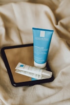 Two skincare products, a blue tube and a white tube, are placed on top of a black-framed mirror, which is resting on crumpled beige fabric.