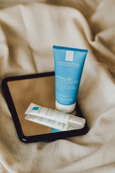 Two skincare products, a blue tube and a white tube, are placed on top of a black-framed mirror, which is resting on crumpled beige fabric.