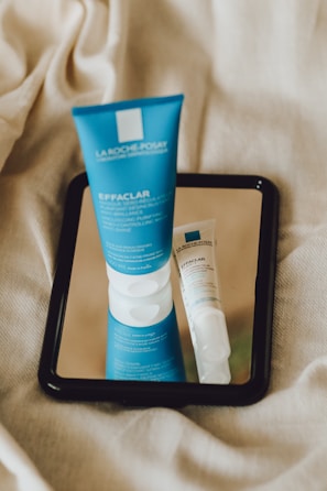 A blue tube of skincare product labeled 'Effaclar' from La Roche-Posay is placed on a mirror, reflecting next to a smaller white and blue tube. The background consists of a beige fabric, giving the setting a soft and natural texture.