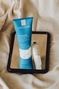 A blue tube of skincare product labeled 'Effaclar' from La Roche-Posay is placed on a mirror, reflecting next to a smaller white and blue tube. The background consists of a beige fabric, giving the setting a soft and natural texture.