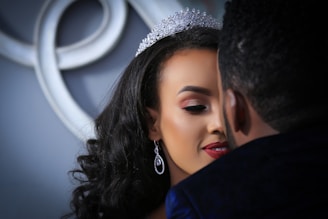 A close-up of a couple embracing, with focus on a woman wearing a sparkling tiara and elegant earrings. Her makeup features bold red lipstick and well-defined eyebrows. The background includes an abstract decorative element.