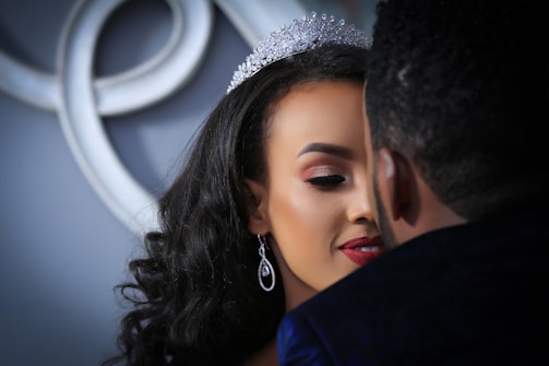 A close-up of a couple embracing, with focus on a woman wearing a sparkling tiara and elegant earrings. Her makeup features bold red lipstick and well-defined eyebrows. The background includes an abstract decorative element.