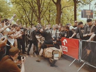 A group of people enthusiastically gather around a vintage motorcycle, which is being started by a rider performing a burnout, evidenced by smoke rising from the rear tire. The crowd appears to be a mix of photographers and spectators, with many holding cameras, and the backdrop is a lush, green park area. A red banner in the background suggests an organized event.
