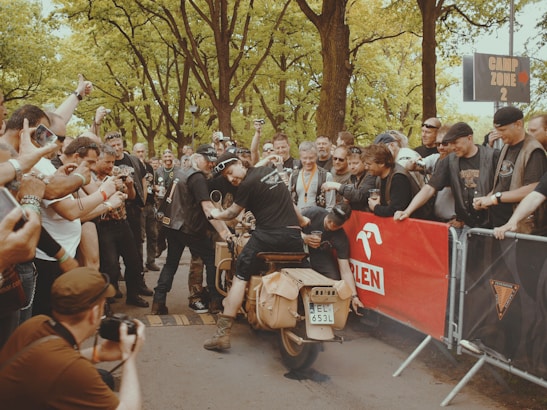 A group of people enthusiastically gather around a vintage motorcycle, which is being started by a rider performing a burnout, evidenced by smoke rising from the rear tire. The crowd appears to be a mix of photographers and spectators, with many holding cameras, and the backdrop is a lush, green park area. A red banner in the background suggests an organized event.