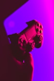 A video game controller is shown in close-up, held in a hand. The lighting is striking, with vivid neon colors of pink and purple that fill the background, creating a dramatic effect.