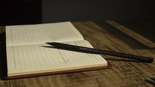 An open notebook with lined pages is resting on a wooden surface, and a black pencil, labeled 'Museo de Arte Latinoamericano Buenos Aires MALBA', is placed diagonally across the notebook.