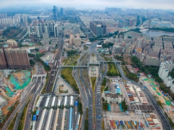 Aerial view of a large urban cityscape with a network of roads, highways, and tall buildings. Skyscrapers dominate the skyline, and the city's infrastructure includes various buildings, bridges, and green areas. In the foreground, there are buildings with colorful rooftops and numerous vehicles on the roads.