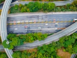 An aerial view of a highway interchange surrounded by lush green trees. Several cars are traveling on multiple lanes, with some vehicles stopped or moving in the opposite direction. Curved overpasses and road markings are visible, indicating a complex road system.