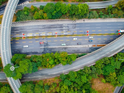 An aerial view of a highway interchange surrounded by lush green trees. Several cars are traveling on multiple lanes, with some vehicles stopped or moving in the opposite direction. Curved overpasses and road markings are visible, indicating a complex road system.