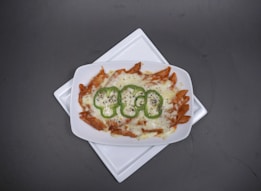 A rectangular white dish contains baked pasta with melted cheese on top, garnished with slices of green bell pepper. The pasta appears to be penne rigate, and there are visible specks of herbs, possibly pepper or oregano, sprinkled over the dish. The dish is centered on a slightly larger white square plate, all placed against a dark gray background.