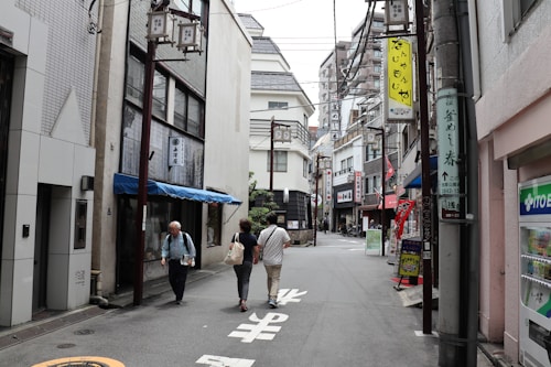 A narrow urban street lined with a variety of small businesses and signs in Japanese script. The buildings feature a mix of modern and traditional architecture, including a blue awning on one of the storefronts. Three people are walking down the street, heading towards the more densely packed area in the background with multi-story buildings.