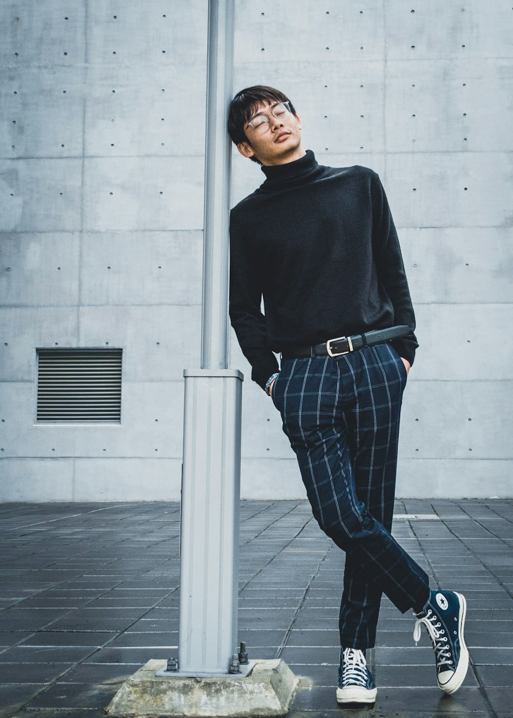 검은 스웨터와 파란색과 흰색 격자 무늬 바지를 입은 남자가 흰색 콘크리트 벽 옆에 서 있습니다.