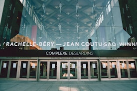 The image depicts the exterior of Complexe Desjardins, featuring a glass facade with reflections of the sky and nearby buildings. Large white text at the top lists store names, while the middle showcases the name Complexe Desjardins. Below, several entrances with glass doors display signs, and the pavement in front is visible.