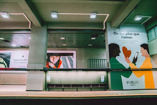 A subway station with concrete pillars and benches is visible, featuring several large advertisements on the walls. One ad has artistic illustrations of two people holding a heart and a smartphone, while another ad showcases a close-up photo of a person's face. The lighting is artificial and the space appears empty of commuters.