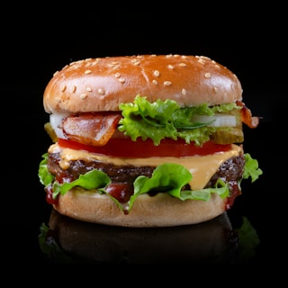 A close-up of a classic hamburger featuring a sesame seed bun, crispy bacon, fresh lettuce, tomatoes, pickles, onions, melted cheese, and a beef patty. The glossy bun and vibrant ingredients create an appetizing presentation.