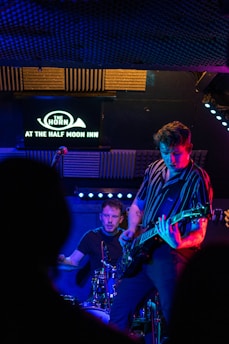 Two musicians perform on a dimly lit stage, one playing the guitar and the other on drums, with vibrant blue and purple lighting. Acoustic panels and a sign reading 'The Horn at The Half Moon Inn' are visible in the background.
