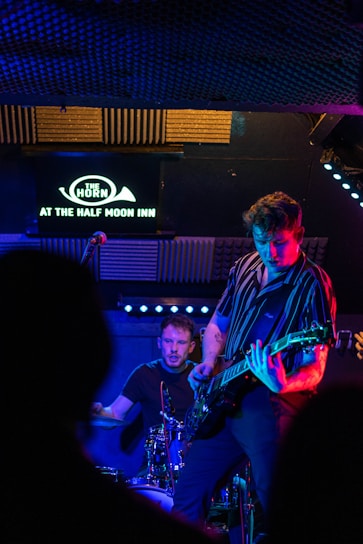 Two musicians perform on a dimly lit stage, one playing the guitar and the other on drums, with vibrant blue and purple lighting. Acoustic panels and a sign reading 'The Horn at The Half Moon Inn' are visible in the background.